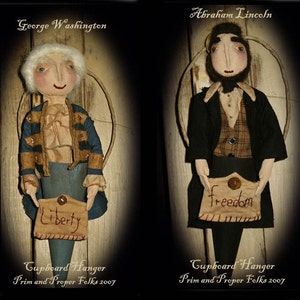 Puede incluir: Dos muñecas de trapo hechas a mano vestidas como figuras históricas, George Washington y Abraham Lincoln. Ambas muñecas sostienen una pequeña bolsa de tela con las palabras "Liberty" y "Freedom" bordadas en ellas. Las muñecas están colgadas de una superficie de madera.
