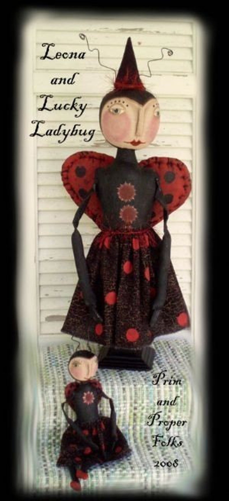 May include: Two handmade rag dolls dressed as ladybugs. The larger doll is standing and has a black dress with red polka dots. The smaller doll is sitting and has a similar dress. Both dolls have black wire arms and legs. The text "Leona and Lucky Ladybug" is written above the dolls. The text "Prim and Proper Folks 2008" is written below the dolls.