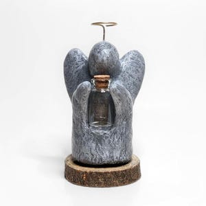 May include: A gray stone angel figurine holding a small glass bottle with a cork stopper. The angel has a halo and is standing on a wooden base.