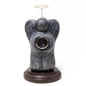 May include: A gray angel figurine with a gold halo and a circular opening. The angel is on a brown, circular base. The figurine has a textured appearance and is set against a white background.