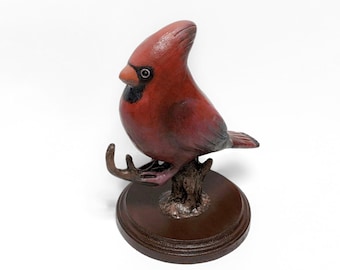 May include: A red cardinal bird figurine perched on a branch, with a detailed painted finish. The bird is sitting on a brown wooden base.