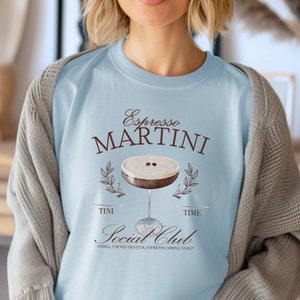 May include: Light blue t-shirt with a brown graphic of an espresso martini cocktail. The text "Espresso Martini" is above the cocktail and "Social Club" is below the cocktail. The text "Tini Time" is on either side of the cocktail. The text "Vodka, Coffee Liqueur, Espresso, Simple Syrup" is below the "Social Club" text.