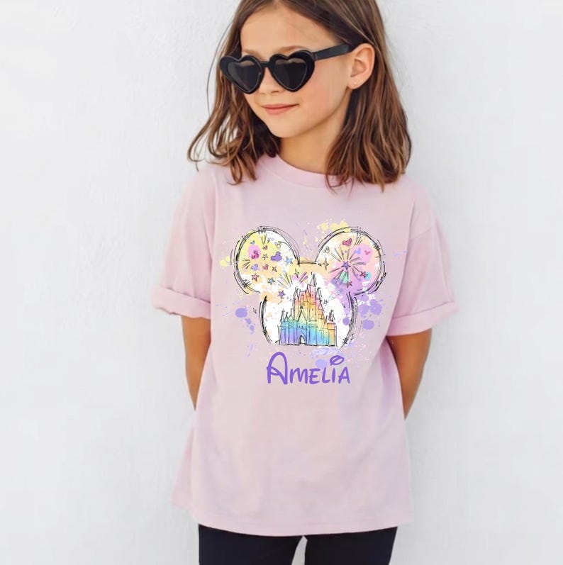 May include: A pale pink t-shirt with a colourful graphic of a castle within Mickey Mouse ears, with fireworks and stars. The name "Amelia" is written in purple script below the graphic. The shirt is worn by a person wearing heart-shaped sunglasses.