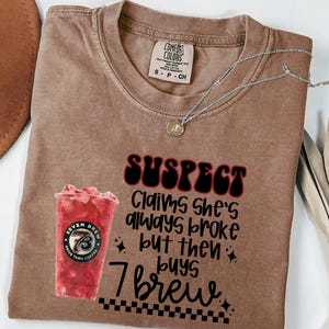 May include: Brown Comfort Colors t-shirt with the text "SUSPECT Claims she's always broke but then buys 7 brew" and a graphic of a red drink. A silver necklace with a pendant is on the shirt.
