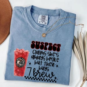 May include: A light blue Comfort Colors t-shirt with the text "SUSPECT Claims she's always broke but then buys 7 Brew." A gold necklace and a Seven Brew coffee cup are also visible.