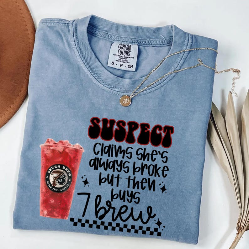 May include: A light blue Comfort Colors t-shirt with the text "SUSPECT Claims she's always broke but then buys 7 Brew." A gold necklace and a Seven Brew coffee cup are also visible.