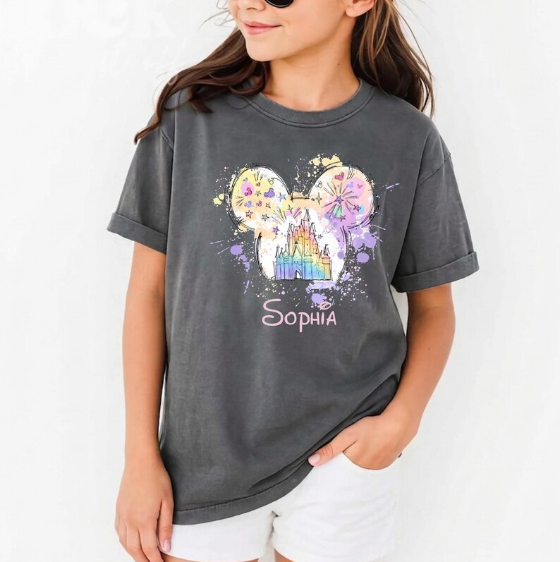May include: Dark grey t-shirt with a colourful graphic of a castle inside Mickey Mouse ears, and the name "Sophia" below. The design includes pastel colours and paint splatters. A casual wear item.