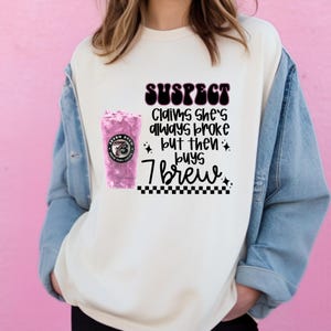 May include: Cream-colored sweatshirt with the text "SUSPECT Claims she's always broke but then buys 7 Brew." A pink 7 Brew drink is on the left side of the graphic. The sweatshirt is worn with a denim jacket.