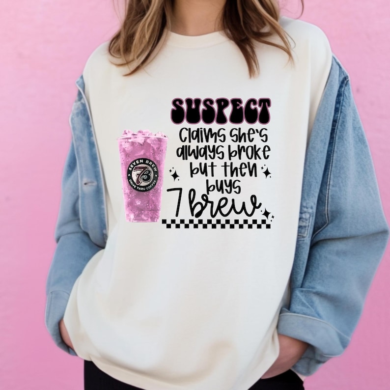 May include: Cream-colored sweatshirt with the text "SUSPECT Claims she's always broke but then buys 7 Brew." A pink 7 Brew drink is on the left side of the graphic. The sweatshirt is worn with a denim jacket.