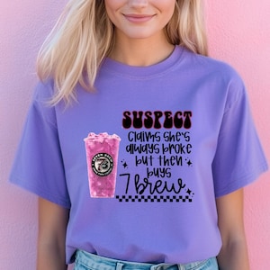 May include: A lilac-colored t-shirt with the text "SUSPECT Claims She's always broke but then buys 7 brew." The graphic includes a pink drink with the Seven Brew logo. The shirt is a casual wear item.