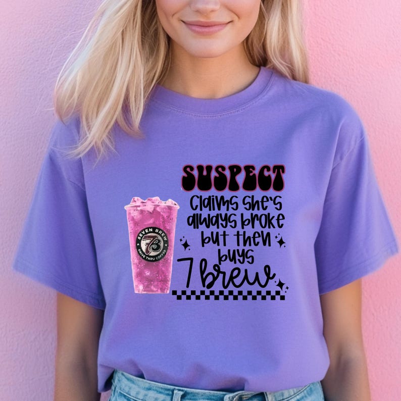 May include: A lilac-colored t-shirt with the text "SUSPECT Claims She's always broke but then buys 7 brew." The graphic includes a pink drink with the Seven Brew logo. The shirt is a casual wear item.