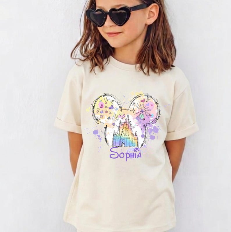 May include: A cream-coloured t-shirt with a colourful graphic of a castle inside a Mickey Mouse head outline. The name "Sophia" is written in purple script below the castle. The design includes fireworks and paint splatters.
