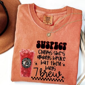 May include: Orange Comfort Colors t-shirt with the text "SUSPECT Claims she's always broke but then buys 7 brew." A graphic of a red drink from Seven Brew is on the left. A gold necklace and a brown hat are also visible.