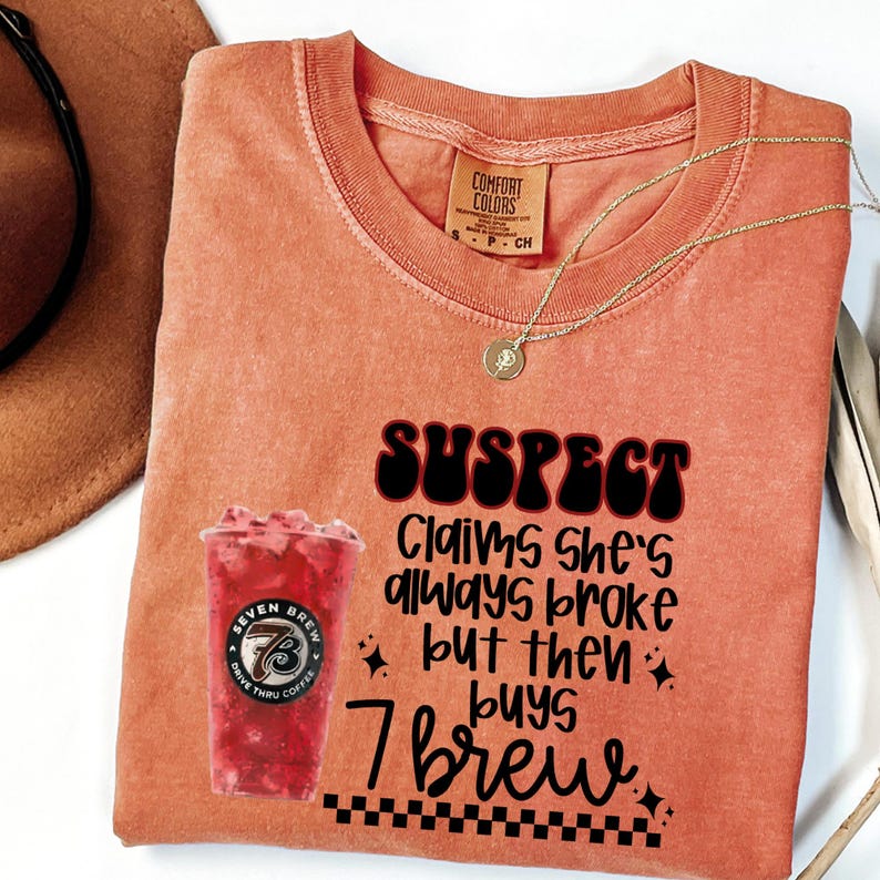 May include: Orange Comfort Colors t-shirt with the text "SUSPECT Claims she's always broke but then buys 7 brew." A graphic of a red drink from Seven Brew is on the left. A gold necklace and a brown hat are also visible.