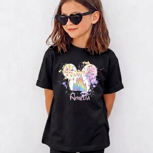 May include: Black t-shirt with a colourful graphic of a castle and fireworks in the shape of Mickey Mouse ears, with the name "Amelia" below. The shirt is worn by a person wearing heart-shaped sunglasses.