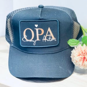 Könnte beinhalten: Schwarze Trucker-Kappe mit einem schwarzen Patch, auf dem "OPA" in roségoldenen Buchstaben mit einem kleinen weißen Herzen über den Buchstaben und "Cindy Anton" in weißer Kursivschrift unter den Buchstaben steht.