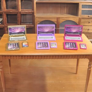 May include: A collection of miniature toy laptops, tablets, and smartphones in gold, purple, and pink. The laptops are open, displaying a screen image. The tablets and smartphones are also visible on a wooden table.