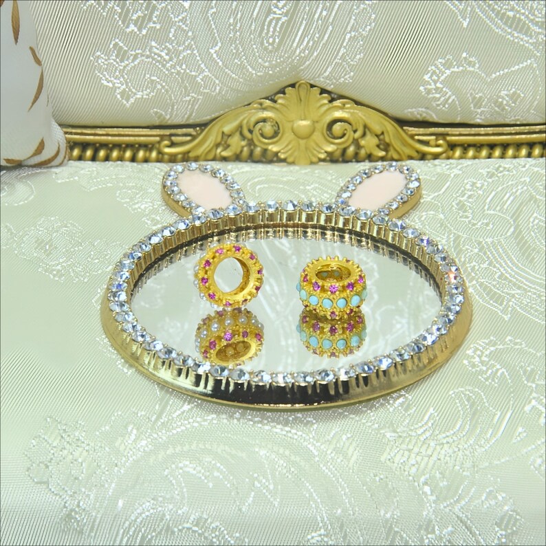 May include: Two gold-toned rings with pink and blue gemstones are displayed on a gold tray with a mirrored surface. The tray is decorated with a white and gold bunny design.