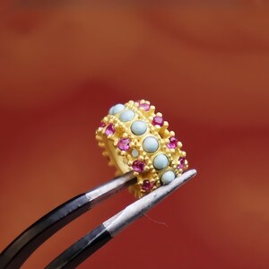 May include: A gold ring with a double row of light blue and pink gemstones. The ring is held by tweezers.