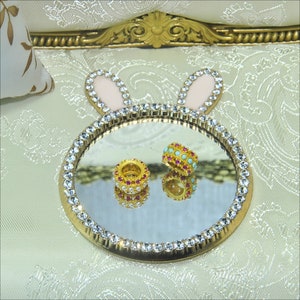 May include: A gold-colored tray with a mirrored surface and a crystal-like border. The tray is shaped like a bunny with pink ears. Two small rings with pink and turquoise stones are on the tray.