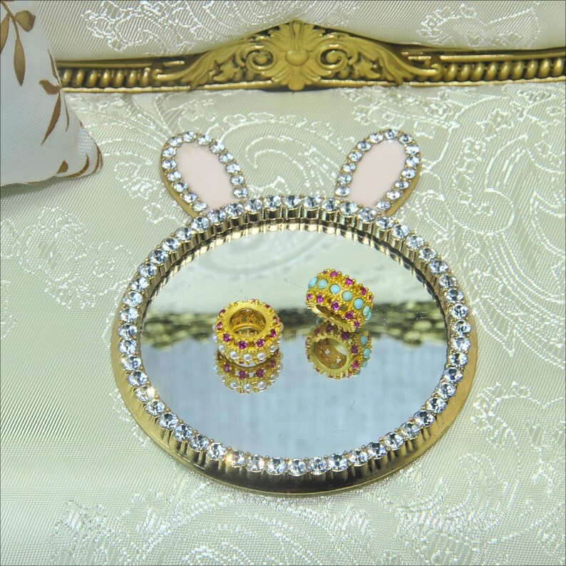 May include: A gold-colored tray with a mirrored surface and a crystal-like border. The tray is shaped like a bunny with pink ears. Two small rings with pink and turquoise stones are on the tray.