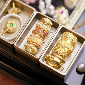 May include: A collection of gold rings with intricate designs and pink and white gemstones. The rings are stacked in a display case with a black background.