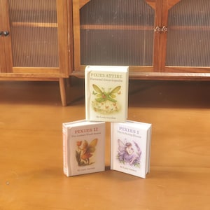 May include: Three miniature books with white covers. The top book is titled "PIXIES ATTIRE Pictorial Encyclopedia" with a fairy illustration. The other two books are titled "PIXIES II The Lanterns Truly Sweet" and "PIXIES I The Echoing Bloom".
