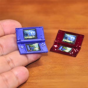 May include: Miniature blue and red handheld gaming consoles, each displaying a colorful game scene on their screens. The consoles are open, showcasing the details of the game graphics and the button layout.