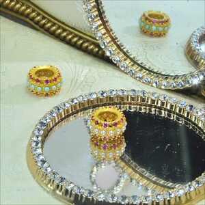 May include: Two gold rings with pink and turquoise stones on a mirrored surface with a gold and crystal trim.