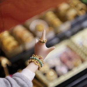 May include: A hand wearing a gold ring with red and green gemstones and a turquoise and gold bracelet with red gemstones. The hand is reaching up towards a blurred background of gold and brown.