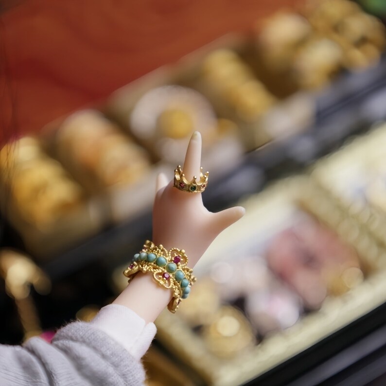 May include: A hand wearing a gold ring with red and green gemstones and a turquoise and gold bracelet with red gemstones. The hand is reaching up towards a blurred background of gold and brown.