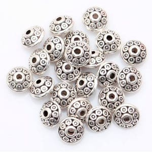 May include: A collection of silver-toned metal beads with a decorative, circular pattern. The beads have a hole in the center for stringing.