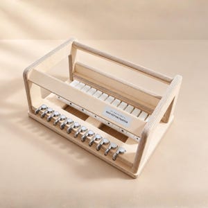 Puede incluir: Un cortador de jabón de madera clara que presenta una serie de perillas de tensión de metal a lo largo del borde frontal y un riel guía blanco con ranuras. Una etiqueta rectangular en el lateral muestra el texto SOAP CUTTER CRAFTER.