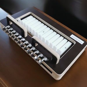 May include: A wooden soap cutter with a black finish and white plastic inserts. The cutter has a handle on the side and a label that reads "SOAP CUTTER CRAFTERS".