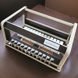 May include: A wooden soap cutter crafter with a brown base and white top. The crafter has a label that reads "Soap Cutter Crafter". The crafter has a series of metal hooks on the bottom and a grid of white squares on the top.
