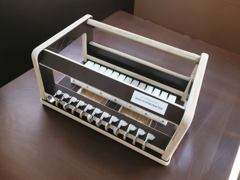 May include: A wooden soap cutter crafter with a brown base and white top. The crafter has a label that reads "Soap Cutter Crafter". The crafter has a series of metal hooks on the bottom and a grid of white squares on the top.