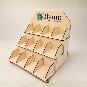 Op de afbeelding: Een displaystandaard met drie niveaus van licht hout, voorzien van verdelers op elke plank om individuele vakken te creëren. Het bovenste achterpaneel toont een groen bloemicoon en de tekst lilyum ethical care in donkerblauwe letters.
