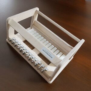 May include: A wooden soap cutter crafter with a white label that reads "Soap Cutter Crafter". The crafter has a series of metal hooks on the bottom and a wooden frame with white bars.
