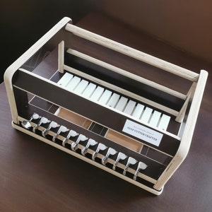 May include: A wooden soap cutter crafter with a white and brown finish. The crafter has a shelf for storing soap cutters and a compartment for storing soap. The text "SOAP CUTTER CRAFTERS" is printed on the front of the crafter.