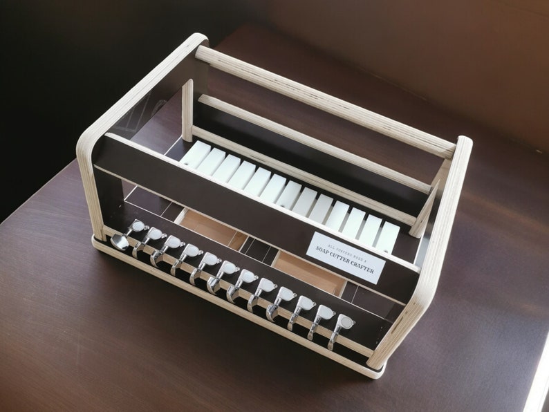 May include: A wooden soap cutter crafter with a white and brown finish. The crafter has a shelf for storing soap cutters and a compartment for storing soap. The text "SOAP CUTTER CRAFTERS" is printed on the front of the crafter.