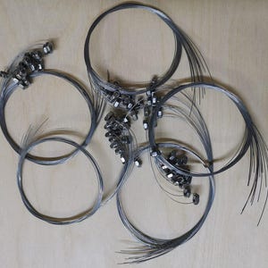 Puede incluir: Vista cenital de varias cuerdas de guitarra de acero enrolladas con tuercas metálicas. Las cuerdas están dispuestas en círculos superpuestos sobre un fondo claro de vetas de madera. Las cuerdas son de color gris oscuro.