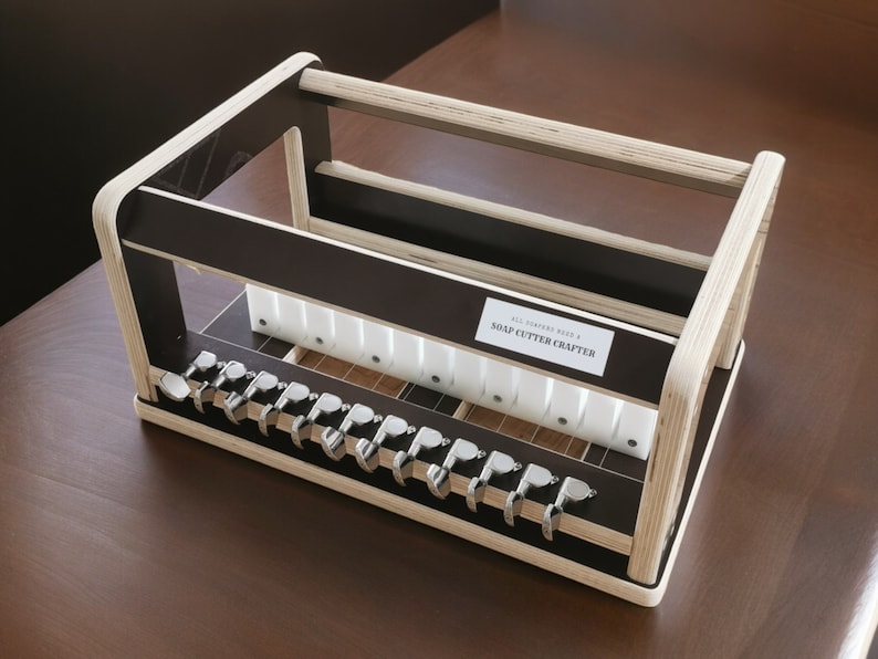 May include: A wooden soap cutter crafter with a black and white design. The crafter has a label that reads "All Natural Reed & Soap Cutter Crafter".