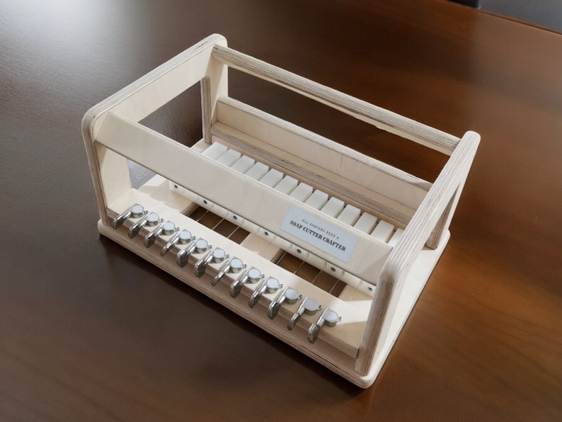May include: A wooden soap cutter crafter with a white label that reads "ALL NATURAL HANDMADE SOAP CUTTER CRAFTERS". The cutter has a series of metal blades and a wooden frame.