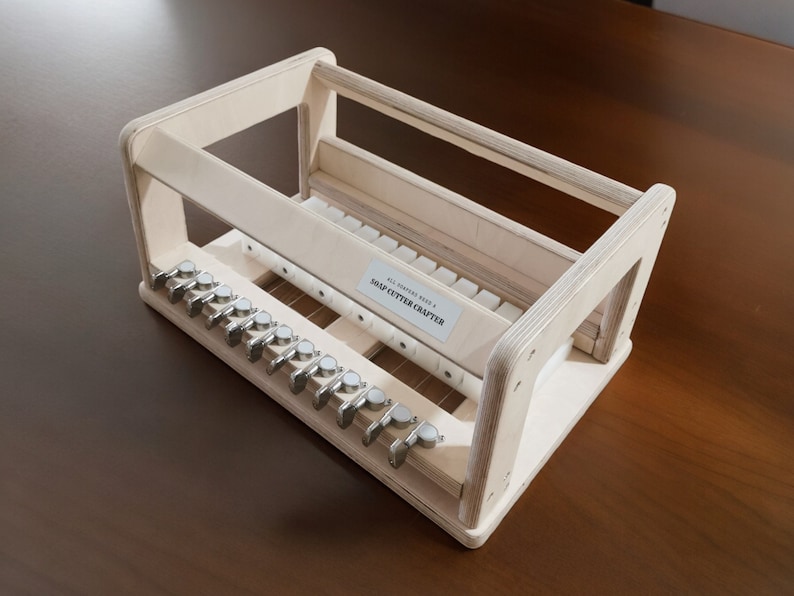 May include: A wooden soap cutter crafter with a white label that reads "ALL NATURAL HANDMADE SOAP CUTTER CRAFTERS". The cutter has a series of metal blades and a wooden frame.