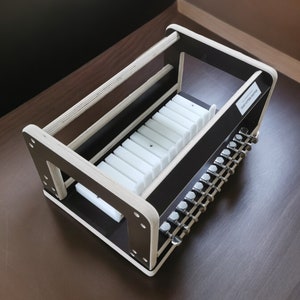 May include: A wooden storage rack with a black base and white dividers. The rack is designed to hold guitar tuning pegs. The rack has a label that reads "Guitar Tuning Pegs".
