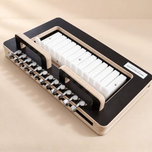 May include: A rectangular soap cutter featuring a black surface with a light wood edge. It includes a white plastic grid insert and a row of silver metal tuning pegs along one side. A small white label on the black surface reads SOAP CUTTER CUTTER.