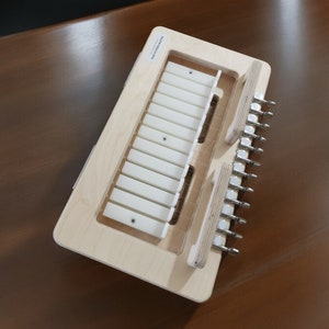 Puede incluir: Una herramienta de madera con una fila de ranuras de plástico blanco y una serie de clavijas metálicas en el lateral. La herramienta está etiquetada como "Guitar Action Gauge".