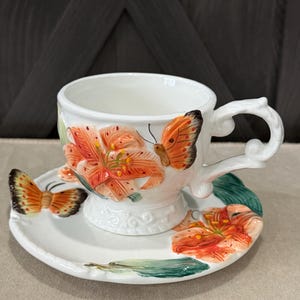 May include: A white ceramic teacup and saucer set featuring three-dimensional orange lily flowers and orange butterflies. The cup has a curved handle and a pedestal base, while the saucer displays a matching floral and butterfly motif in relief.