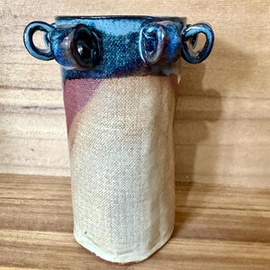 May include: A cylindrical ceramic vase featuring a textured, off-white body with a subtle pinkish-tan diagonal band. The upper rim is finished in a deep, glossy blue glaze and is adorned with four small, looped handles extending outward.