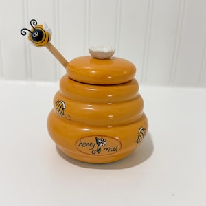 May include: Orange ceramic honey pot with a lid and a wooden honey dipper. The pot is shaped like a beehive and decorated with painted bees and the words "honey miel". The honey dipper has a bee-shaped handle.